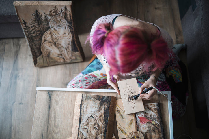 Person with pink hair drawing a picture of a cat on a wooden plaque.